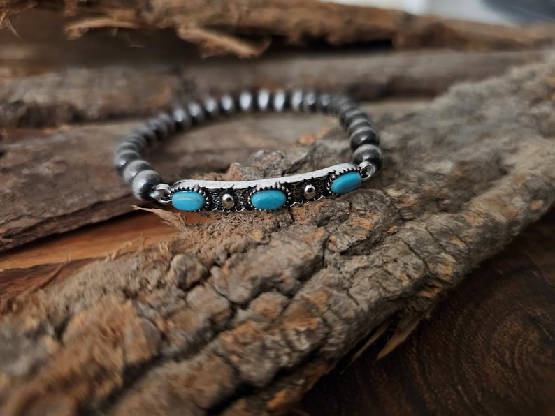 May include: A beaded bracelet with a silver bar featuring three oval turquoise stones. The bracelet is made of small, round, silver-colored beads. The jewelry is displayed on a piece of textured, brown wood.