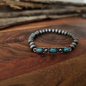 May include: A beaded bracelet with a silver accent featuring three oval turquoise stones. The bracelet is made of small, gray beads and is displayed on a wooden surface.