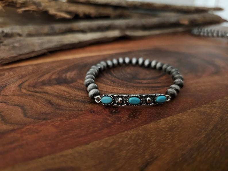 May include: A beaded bracelet with a silver accent featuring three oval turquoise stones. The bracelet is made of small, gray beads and is displayed on a wooden surface.