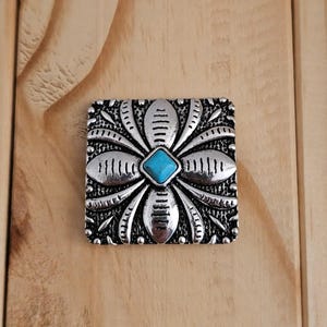 May include: A silver-toned, square-shaped decorative embellishment with a floral design. The center features a small, square turquoise-colored stone. The embellishment is set against a wooden background.