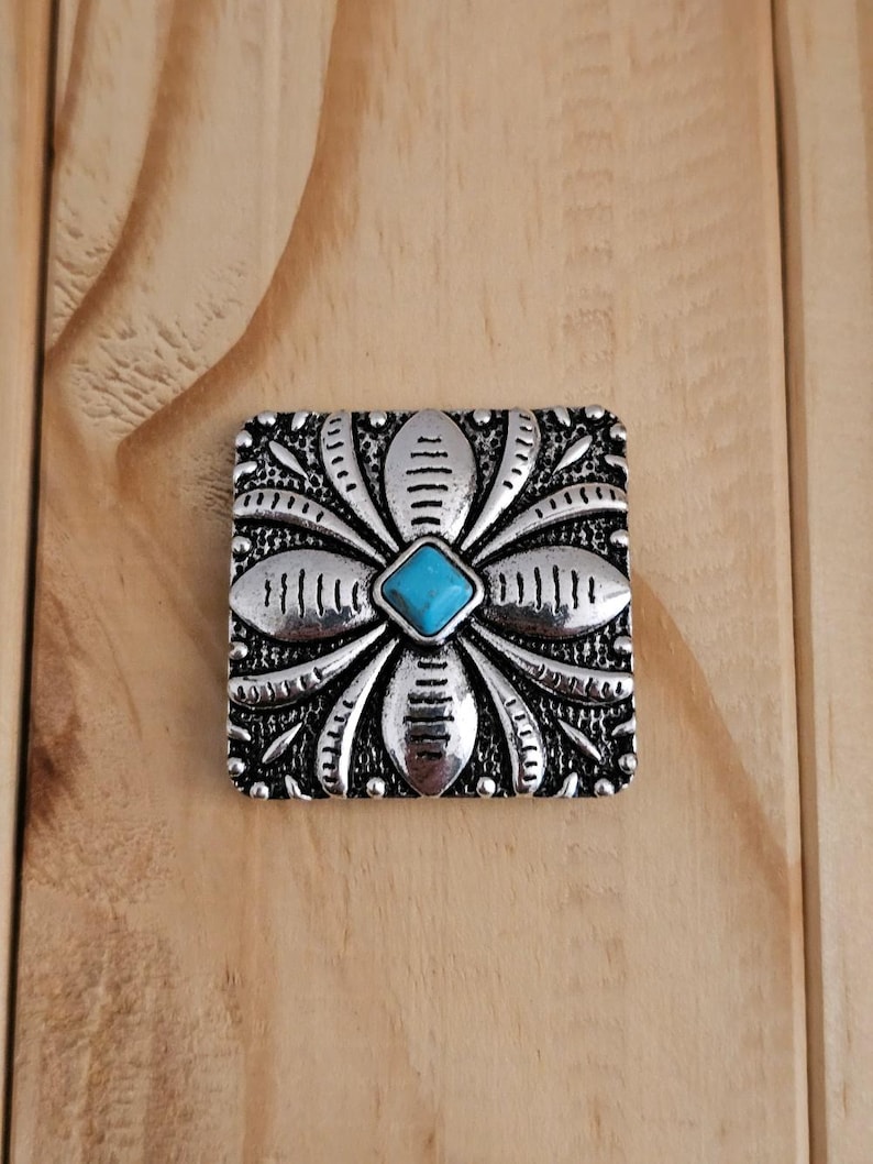 May include: A silver-toned, square-shaped decorative embellishment with a floral design. The center features a small, square turquoise-colored stone. The embellishment is set against a wooden background.