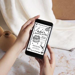 May include: A smartphone displays a black and white invitation for "Katie's Divorce Party." The invitation includes a cake, disco ball, and martini glass illustrations. The phone is held in front of a neutral background with two vases.