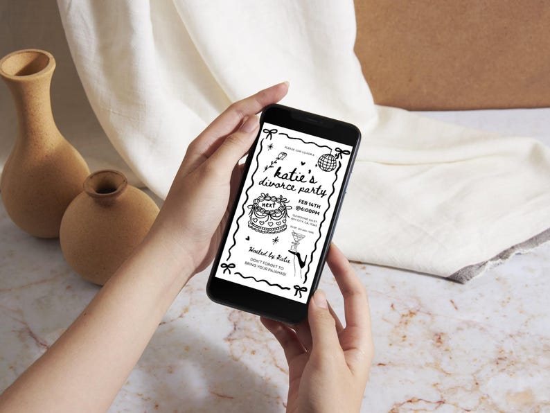 May include: A smartphone displays a black and white invitation for "Katie's Divorce Party." The invitation includes a cake, disco ball, and martini glass illustrations. The phone is held in front of a neutral background with two vases.