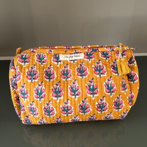 May include: Mustard-colored quilted cosmetic bag with a floral pattern in pink and teal. The bag has a gold zipper and a yellow tassel. The text "Simply Soleil" is printed on a small tag.