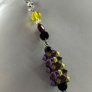 May include: A handmade charm featuring a combination of beads and folded ribbon. The charm includes yellow, maroon, and black beads, along with a section of purple and gold ribbon. It is attached to a silver-colored clasp and a lavender cord.