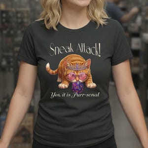 以下が含まれることがあります： ダークグレーのTシャツ。ティアラとピンクのメガネをかけたオレンジ色の漫画の猫が、紫色の20面ダイスを持っています。「Sneak Attack!」と「Yes, it is Purr-sonal」の文字が白いスクリプトで表示されています。