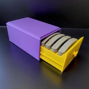 May include: A purple rectangular box with a yellow drawer, containing four gray game cartridges. The drawer has a small, round yellow knob. The box is designed to store video game cartridges.