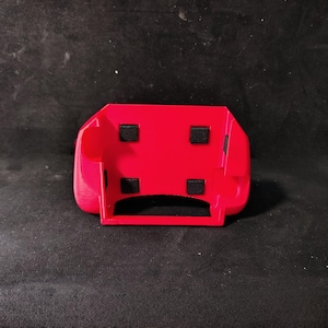 May include: A bright red plastic object with a rectangular shape and rounded edges. Four small, black, square pads are affixed to the top surface. The object has a curved, open-ended design, suggesting it may be a holder or mount.