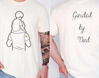 May include: Cream-colored t-shirt featuring a black line drawing of a person carrying a child on their shoulders. The back of the shirt has the words "Guided by Dad" in a cursive font. A small colorful design is on the left shoulder.
