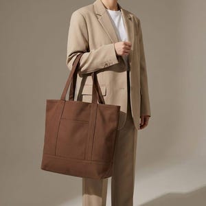 K&ouml;nnte beinhalten: Eine braune Tragetasche mit rechteckiger Form und einer Vordertasche. Die Tasche hat zwei Griffe und wird von einer Person getragen, die einen beigefarbenen Anzug und ein wei&szlig;es T-Shirt tr&auml;gt.