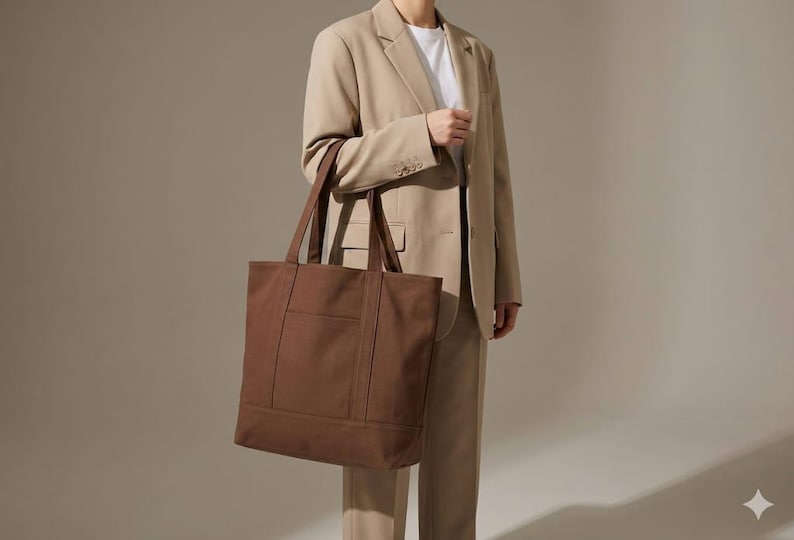 K&ouml;nnte beinhalten: Eine braune Tragetasche mit rechteckiger Form und einer Vordertasche. Die Tasche hat zwei Griffe und wird von einer Person getragen, die einen beigefarbenen Anzug und ein wei&szlig;es T-Shirt tr&auml;gt.
