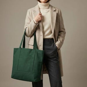 K&ouml;nnte beinhalten: Eine dunkelgr&uuml;ne Tragetasche mit rechteckiger Form und einer Vordertasche. Die Tasche hat zwei Henkel und wird von einer Person getragen, die einen beigefarbenen Mantel, einen cremefarbenen Rollkragenpullover und eine dunkelgraue Hose tr&auml;gt.