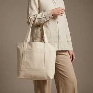 K&ouml;nnte beinhalten: Eine beigefarbene Canvas-Tragetasche mit rechteckiger Form und einer Vordertasche. Die Tasche hat zwei Griffe und wird von einer Person getragen, die ein beiges Hemd und eine Hose tr&auml;gt.