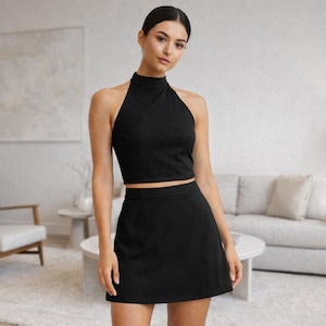 Puede incluir: Conjunto negro de top halter y falda a juego. El top tiene un cuello alto y es sin mangas. La falda es una minifalda. El atuendo lo lleva una persona en un salón moderno y minimalista.