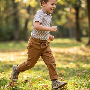 Könnte beinhalten: Ein Kind rennt durch einen Park. Das Kind trägt ein gestreiftes Hemd, eine braune Hose und Turnschuhe. Der Hintergrund ist ein verschwommener grüner Rasen und Bäume, mit Sonnenlicht, das durch die Blätter scheint.