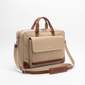 May include: Tan canvas briefcase with brown leather accents. Features a front flap pocket, top handles, and a detachable shoulder strap. The bag has a zipper closure and metal hardware.