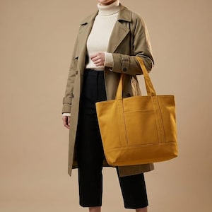 K&ouml;nnte beinhalten: Eine Person tr&auml;gt einen beigefarbenen Trenchcoat, eine schwarze Hose und einen cremefarbenen Rollkragenpullover und h&auml;lt eine senfgelbe Tragetasche. Die Tragetasche hat eine rechteckige Form mit einer Vordertasche und zwei Griffen.