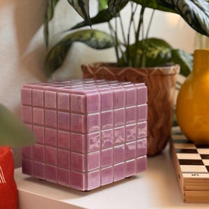 May include: A lavender-coloured cube-shaped decorative object with a tiled surface. The item is made of small, square tiles and sits on a white surface. The object is next to a chess set and a yellow vase.