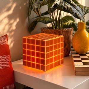Może przedstawiać: Kwadratowa lampa w kształcie kostki logicznej z siatką błyszczących, pomarańczowych kwadratowych płytek. Lampa emituje od wewnątrz ciepłe światło, które podświetla szczeliny między poszczególnymi płytkami. Stoi na płaskiej powierzchni jako dekoracja.