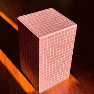 May include: A rectangular pedestal covered in small, square, light pink tiles. The structure features a grid pattern created by the grout lines between each individual tile, forming a uniform, geometric surface on the top and sides of the object.