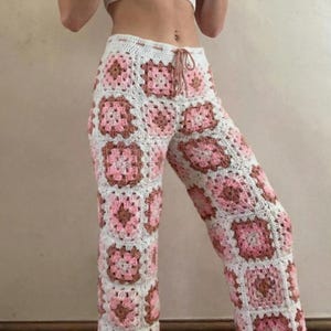 May include: Hand-crocheted pants featuring a patchwork design of white, pink, and brown squares. The pants have a drawstring waist and a wide-leg fit. The crochet work is intricate, creating a textured and visually appealing garment.