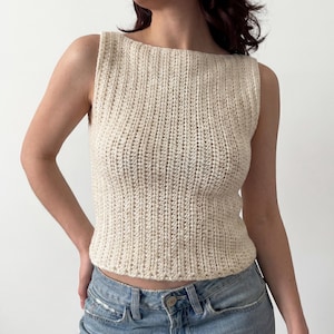 May include: Cream-colored, sleeveless crochet top with a ribbed texture. The top has a boat neck and a fitted silhouette. The yarn appears to be a natural fiber. The top is paired with blue jeans.