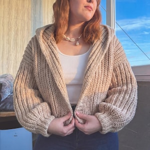 Puede incluir: Un cárdigan beige con capucha y punto grueso. El suéter abierto tiene un patrón de punto grueso y texturizado. Las mangas son largas y la capucha es grande. El cárdigan es de color neutro, adecuado para combinar. La modelo lleva una camiseta sin mangas blanca y un collar de perlas.