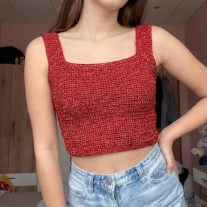 May include: A sleeveless, cropped top made of red yarn with a metallic shimmer. The garment features a square neckline and wide shoulder straps, constructed with a dense, textured crochet stitch pattern that covers the entire torso area.