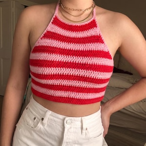 May include: A handmade, red and pink striped crochet halter top. The top features horizontal stripes of alternating colours. The halter top is worn with white shorts and gold necklaces.