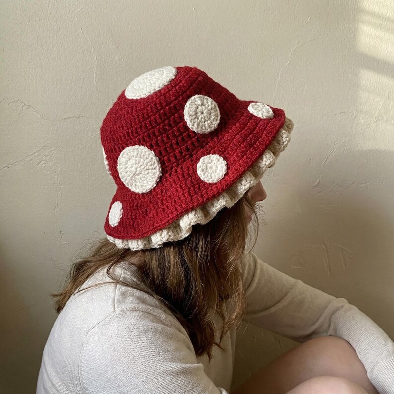May include: A red crochet mushroom hat with white polka dots and a cream-colored ruffled brim. The hat is handmade and has a whimsical, playful design. The hat is a unique accessory.