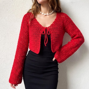 May include: A bright red, open-front crochet cardigan with long sleeves. The cardigan has a tie closure at the bust and a scalloped neckline. The garment is worn over a black dress.
