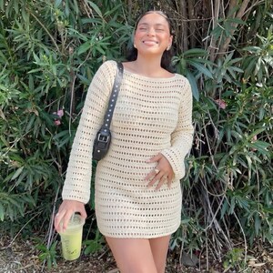 May include: A cream-colored, long-sleeved crochet dress with a scoop neckline. The dress has a loose, open weave design. A black shoulder bag with silver studs is worn across the body. The person is holding a green beverage in a clear plastic cup.