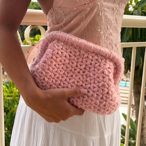 May include: A light pink crochet clutch bag featuring a textured star-like stitch pattern throughout the body. The rectangular pouch has a thick, reinforced top edge and a soft, yarn-based construction with a consistent pastel pink color tone.