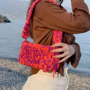 May include: A handmade, vibrant shoulder bag in a striking combination of orange and purple. The bag features a woven design, a matching strap, and a decorative flower. The bag is a stylish accessory for carrying essentials.