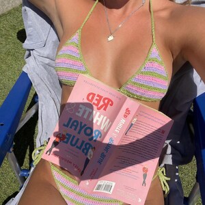 May include: A crochet bikini set featuring horizontal stripes in light green and lavender. The top is a triangle style with thin straps, and the bottom has side ties. A book titled Red, White & Royal Blue is held in front of the bikini.