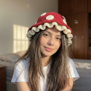 May include: A handmade crochet mushroom hat. The hat features a red top with white polka dots, a cream-colored ruffled brim, and a rounded crown. The hat is designed to be worn as a fashion accessory.