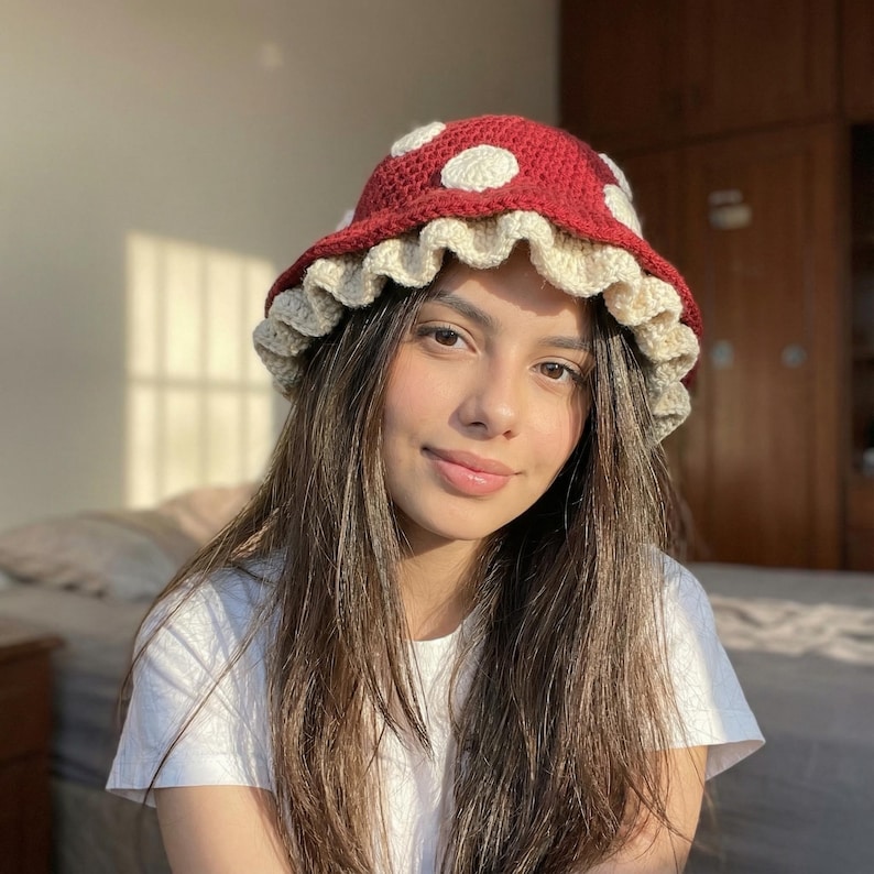 May include: A handmade crochet mushroom hat. The hat features a red top with white polka dots, a cream-colored ruffled brim, and a rounded crown. The hat is designed to be worn as a fashion accessory.