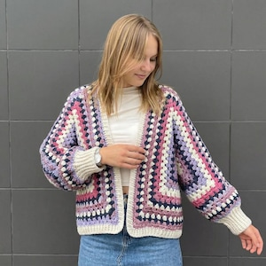 May include: A colorful, open-front crochet cardigan in a granny square pattern. The sweater features a mix of pink, purple, white, and navy blue squares. The sleeves are long, and the cuffs and trim are a cream color.