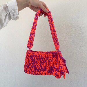May include: A handmade, crochet handbag in vibrant orange and purple. The bag features a rectangular body with a braided strap and a decorative flower. The color combination creates a bold, eye-catching accessory.