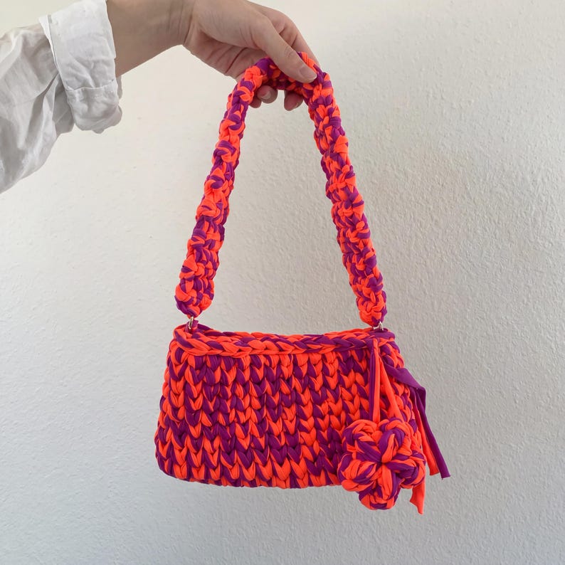 May include: A handmade, crochet handbag in vibrant orange and purple. The bag features a rectangular body with a braided strap and a decorative flower. The color combination creates a bold, eye-catching accessory.