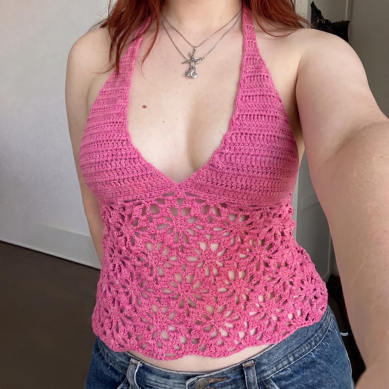 May include: A bright pink crochet halter top featuring a solid stitch triangular bodice and a lower section crafted with an intricate, openwork floral pattern. The garment has a deep V-neckline and ties around the neck, paired with blue denim jeans.