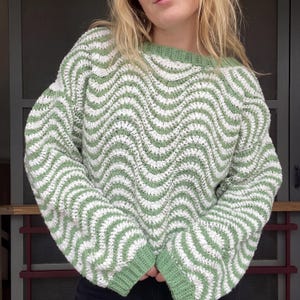 May include: A long-sleeved crochet jumper featuring a repeating wavy pattern in sage green and white. The garment has a round neckline and ribbed cuffs, both finished in a solid sage green yarn, with a loose fit and textured stitch construction.