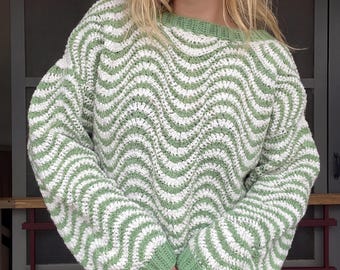 May include: A long-sleeved crochet sweater featuring a repeating wavy pattern in sage green and white. The garment has a round neckline and ribbed cuffs, both finished in a solid sage green yarn, with a loose fit and textured stitch construction.