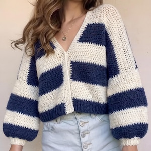 Könnte beinhalten: Handgefertigte Häkeljacke im maritimen Stil. Der Pullover hat abwechselnde horizontale Streifen in Marineblau und Creme. Die Jacke hat einen V-Ausschnitt, lange Ärmel und eine Knopfleiste. Die Ärmel sind an den Schultern leicht aufgebauscht.