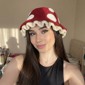 Könnte beinhalten: Häkel-Pilzhut in Rot mit weißen Punkten und einem cremefarbenen, gerüschten Rand. Dieses handgefertigte Accessoire ist ideal, um jedem Outfit eine verspielte Note zu verleihen. Ein einzigartiges, skurriles Design.