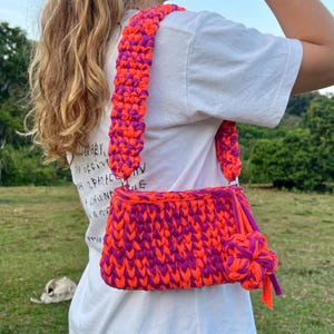 May include: A handmade crochet bag in vibrant orange and purple. The bag features a rectangular body with a matching strap and a decorative flower accent. The bag is made of thick, textured yarn.