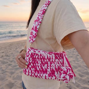 May include: A pink and white crocheted shoulder bag with a matching strap. The bag has a rectangular shape with a tassel and a decorative flower. The bag is made of thick yarn and has gold-colored hardware.