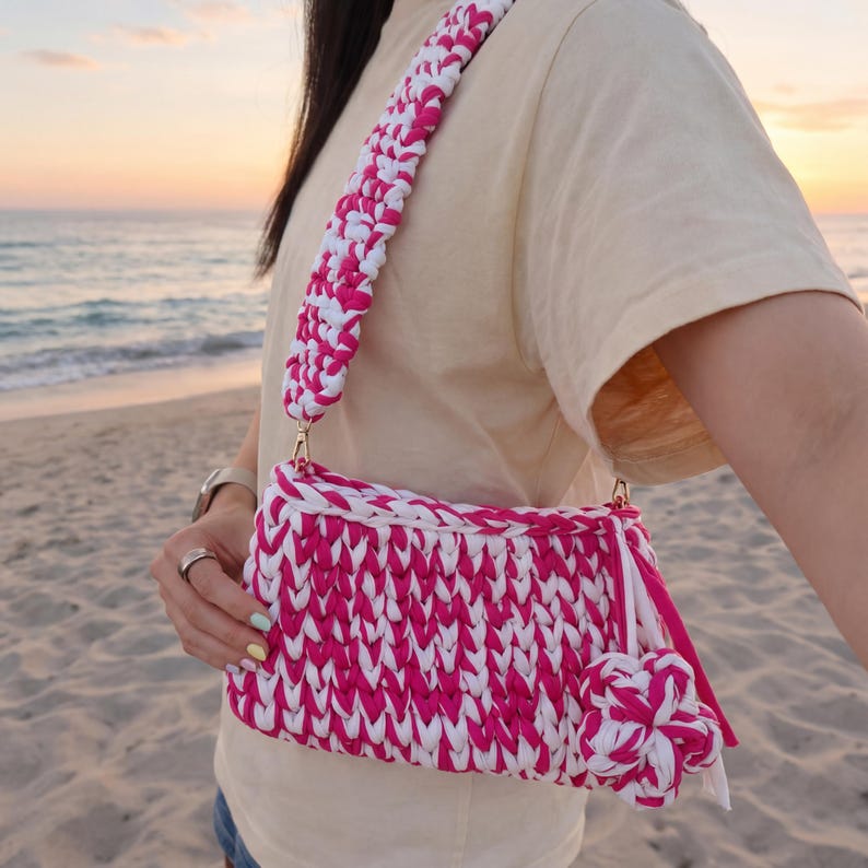May include: A pink and white crocheted shoulder bag with a matching strap. The bag has a rectangular shape with a tassel and a decorative flower. The bag is made of thick yarn and has gold-colored hardware.