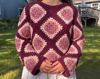 May include: A long-sleeved crochet sweater featuring a repeating granny square pattern in shades of deep burgundy, dusty rose, and cream. The garment has a crew neckline and a boxy fit, constructed from interconnected yarn squares in a textured design.