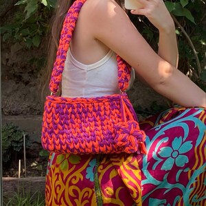May include: A handmade, vibrant crochet bag in shades of orange and purple. The bag features a rectangular body with a matching strap and a decorative knotted detail. The bag is held by a person wearing a white tank top and a colorful skirt.